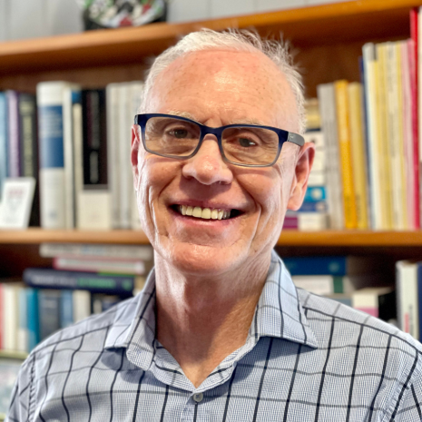 Photo of a person wearing black glasses and a blue check shirt, in front of a book case, smiling at the camera.