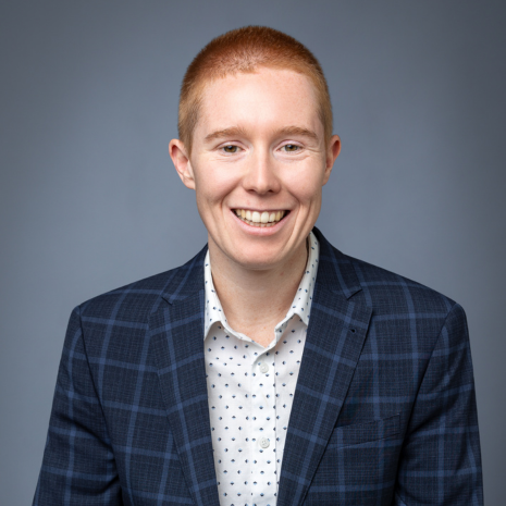 Photo of a person smiling at the camera, wearing a white shirt with polkadots and a navy jacket with check pattern on it.