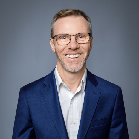 Photo of a person with glasses and short hair, dressed in a shirt with a blazer, smiling at the camera