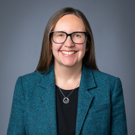 Photo of a person in a teal blazer, with a black shirt and glasses, smiling directly at the camera