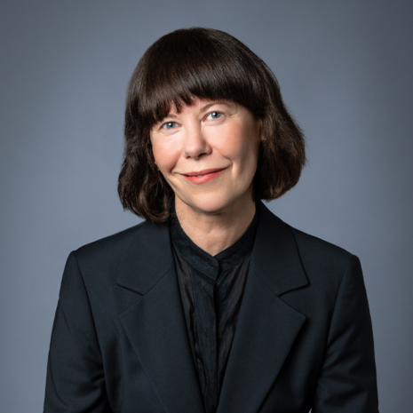 Photograph of a person in a black blazer and brown hair smiling at the camera in front of a light grey background