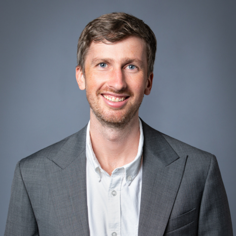 Photo of a person with short hair, smiling at the camera, wearing a grey jacket, collared shirt and in front of a grey background