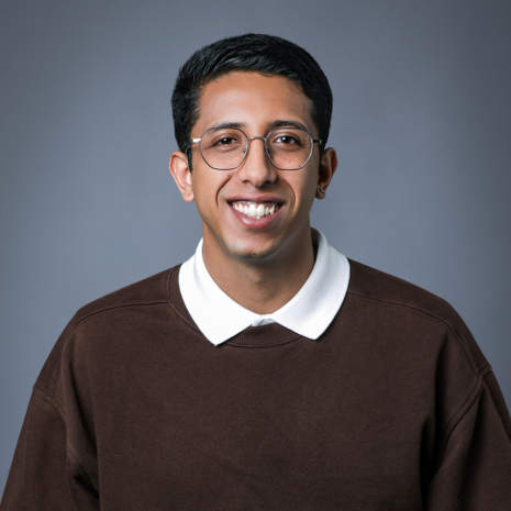 A person with glasses and wearing a white shirt beneath a brown long-sleeved jumper is looking at the camera and smiling.