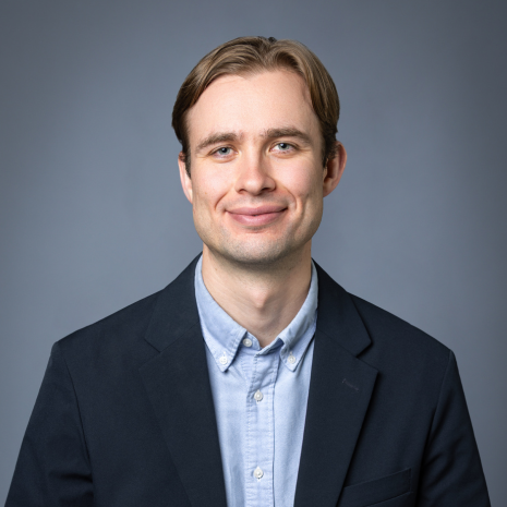Photo of a person in a light blue shirt with a navy blue jacket, looking directly at the camera and smiling. They are sitting in front of a plain grey background.