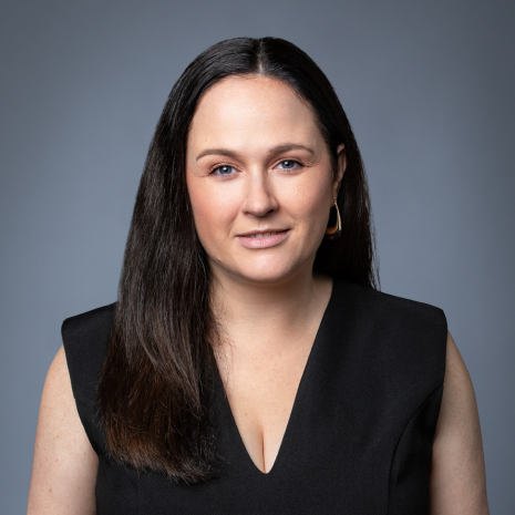 Photo of a person looking directly at the camera and smiling. They are wearing a black top and sitting in front of a grey background.