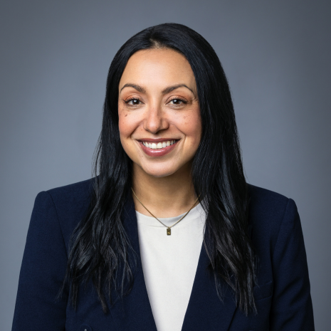 Photo of a person with long dark hair smiling at the camera. They are wearing a white shirt and a dark suit jacket.