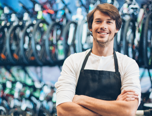 Photo of a small business owner in front of rows of bicycles. They are wearing an apron, folding their arms and smiling at the camera.
