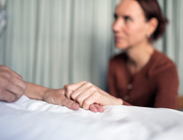 Photo of someone sitting next to a bed, holding the hand of the person in that bed, resting atop a white sheet