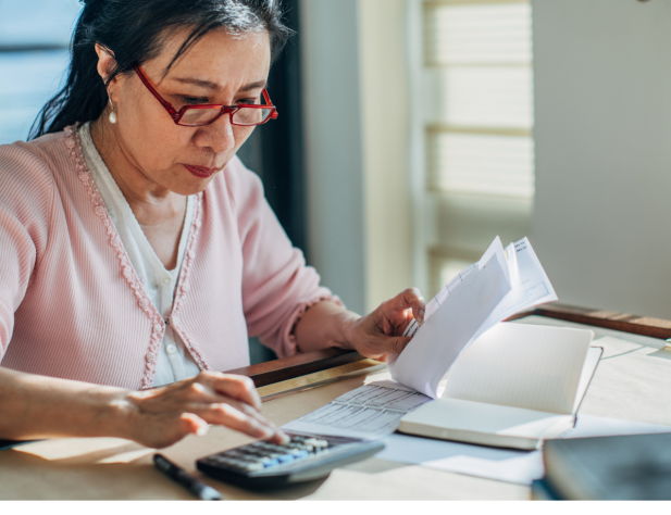 Photo of a person at a desk in part sunlight, wearing glasses and holding paper in their left hand, and tapping a calculator in their right. They are looking at the paper.