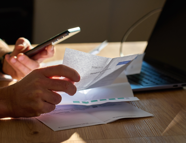 Photo of hands at a desk - one hand is holding a phone and the other is sorting through receipts/bills. There is also a laptop atop the desk.