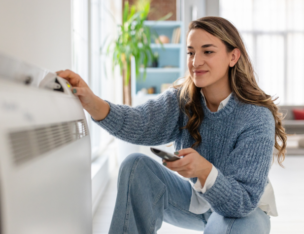 Photo of a woman crouching next to an air conditioner in a home interior, holding remote in her hand and smiling