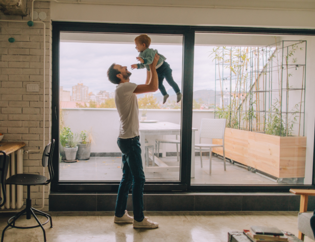 Photo of an interior apartment space, in which a parent is holding their child up in the air in a lounge room setting. Behind them, there is a sliding glass dooe leading to an external balcony area, with some potted plants and a seating area.