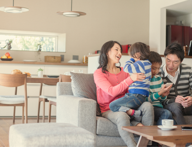 Photo of a family with two parents and two kids, sitting on a lounge in an interior living space. A dining table with chairs, a kitchen, some lighting and a window can be seen in the background.