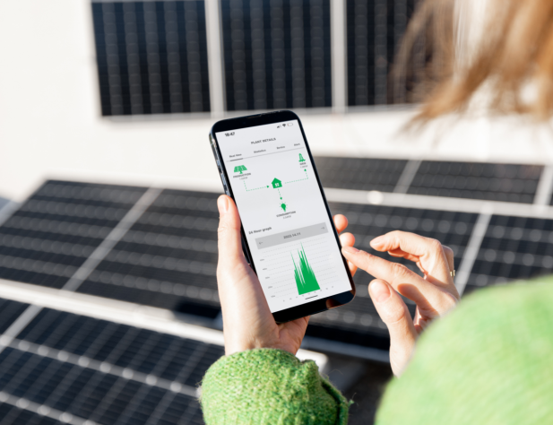 Photo of a person using an energy app, facing away from the camera. They are wearing a green fluffy jumper and standing in front of solar panels. Their hands, holding a phone, and some of their hair is in shot.