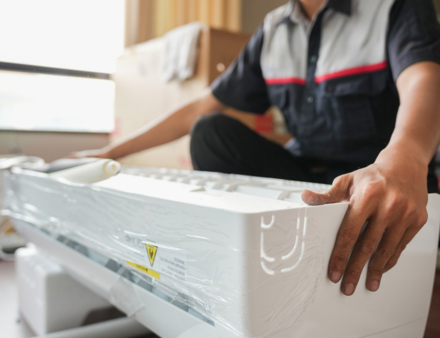 Photo of a person unpacking a new air conditioning unit, while seated on the ground in an interior setting.