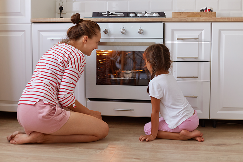 Photo of a woman and child peering into an oven as something bakes. They are seated on the floor of a kitchen, surrounded by white cabinetry.