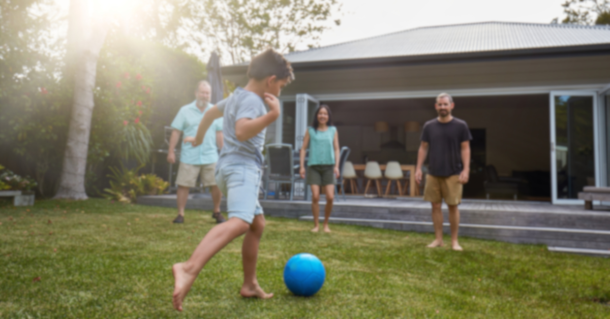 Photo of a kid kicking a blue ball in their backyard with their family watching on, in front of their house